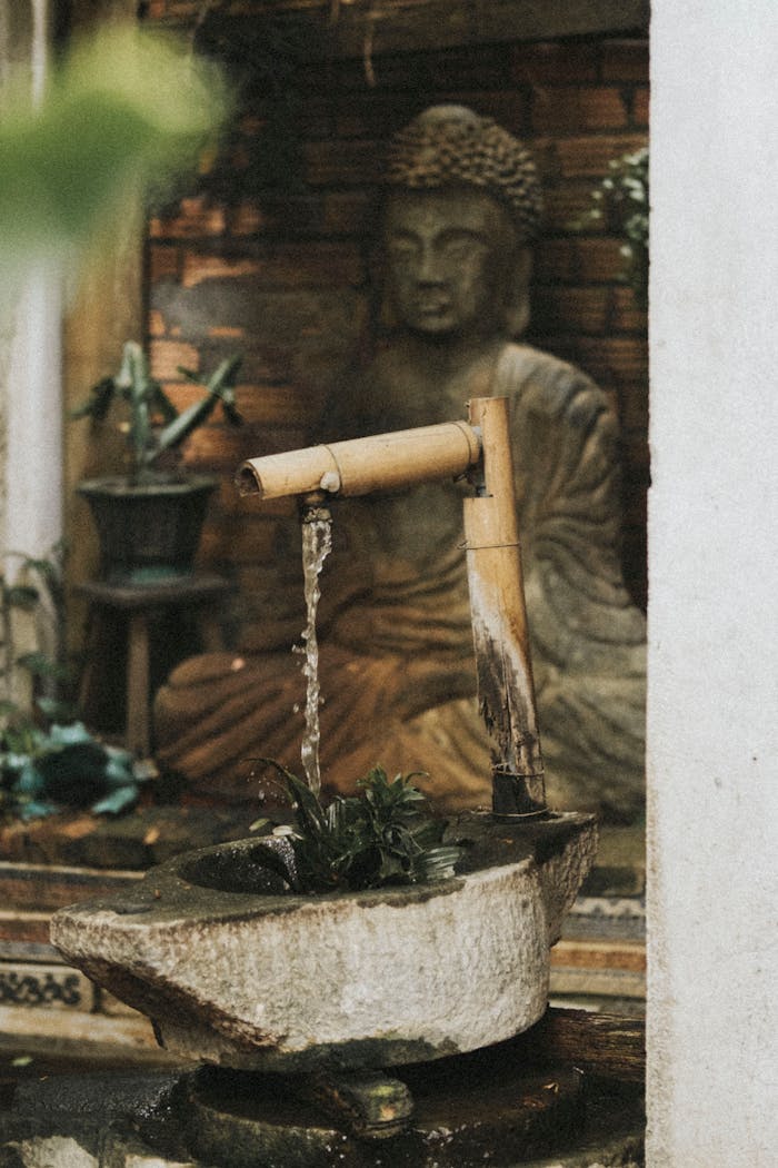 about-bg Tranquil scene of a bamboo water fountain beside a Buddha statue in a garden setting.