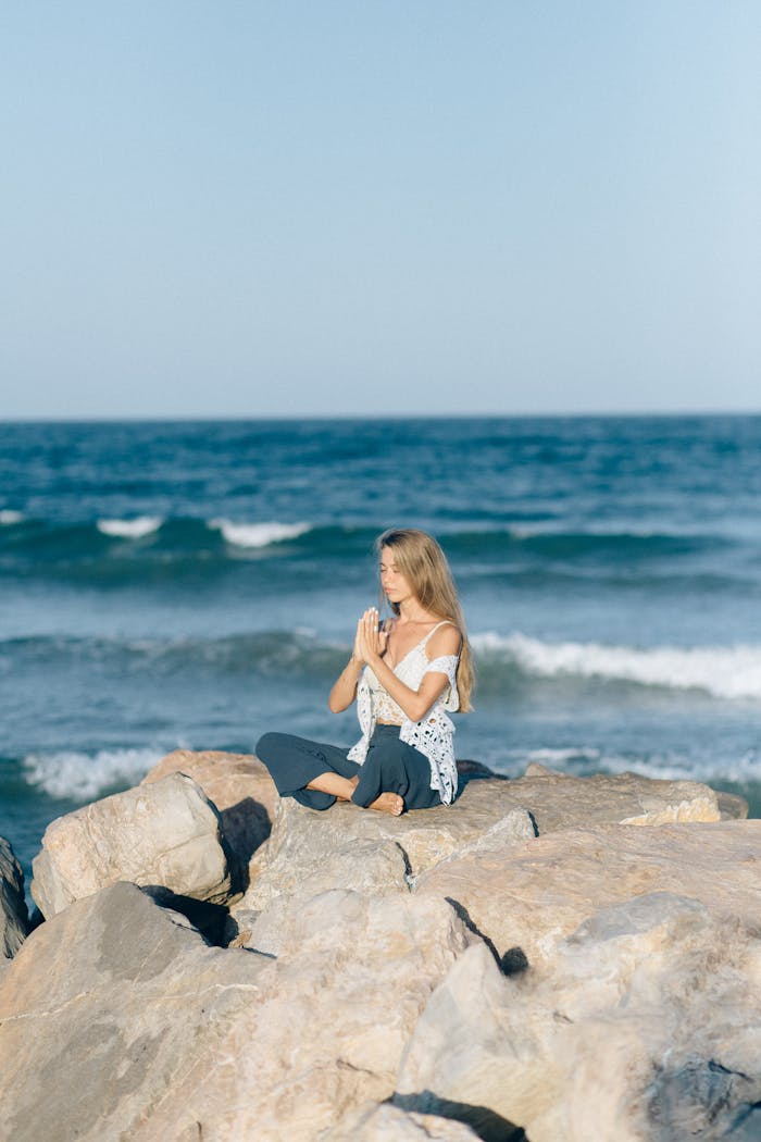 Woman meditating on rocky beach with calm sea in background.